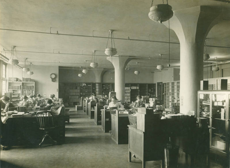 1913 Catalog Department. Workers sitting at desks in a large room with shelves of books around the parameter.