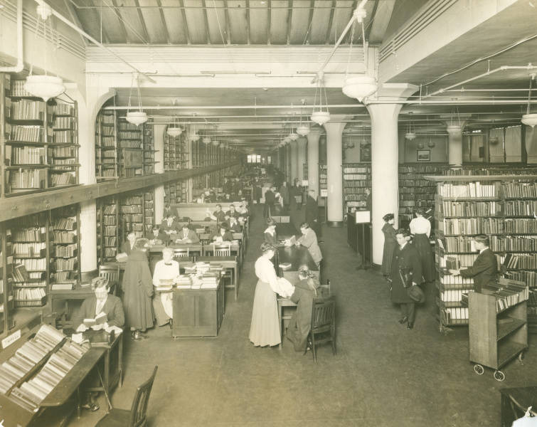 Long room filled with shelved books, along the sides and a filled with patrons sitting at desks reading