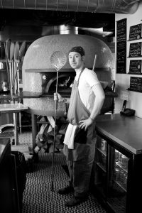 Picture of a man in t-shrit and jeans, standing in front of a wood fire oven