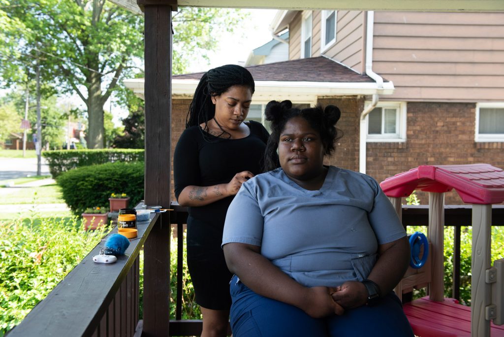 A woman braiding another woman's hair while sitting on a shaded porch in the Collinwood Neighborhood.
