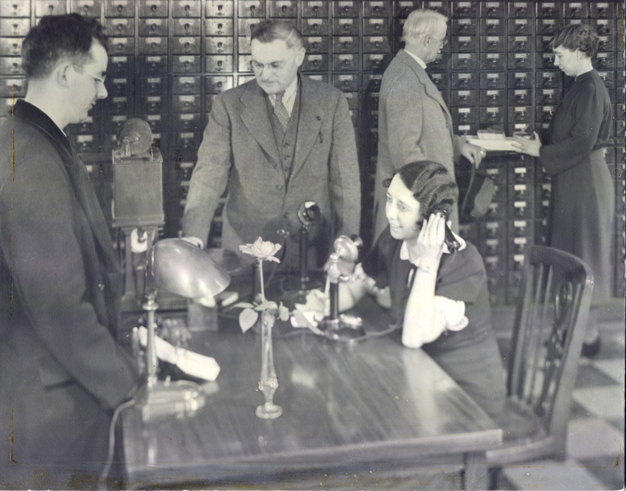 Black and white of female librarian on the phone at a table in front of 3 men and the wall-size card catalog.