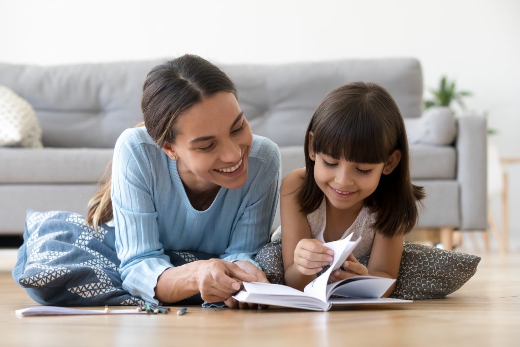 Caring smiling mother reading book with little kid girl lying on warm floor at home.