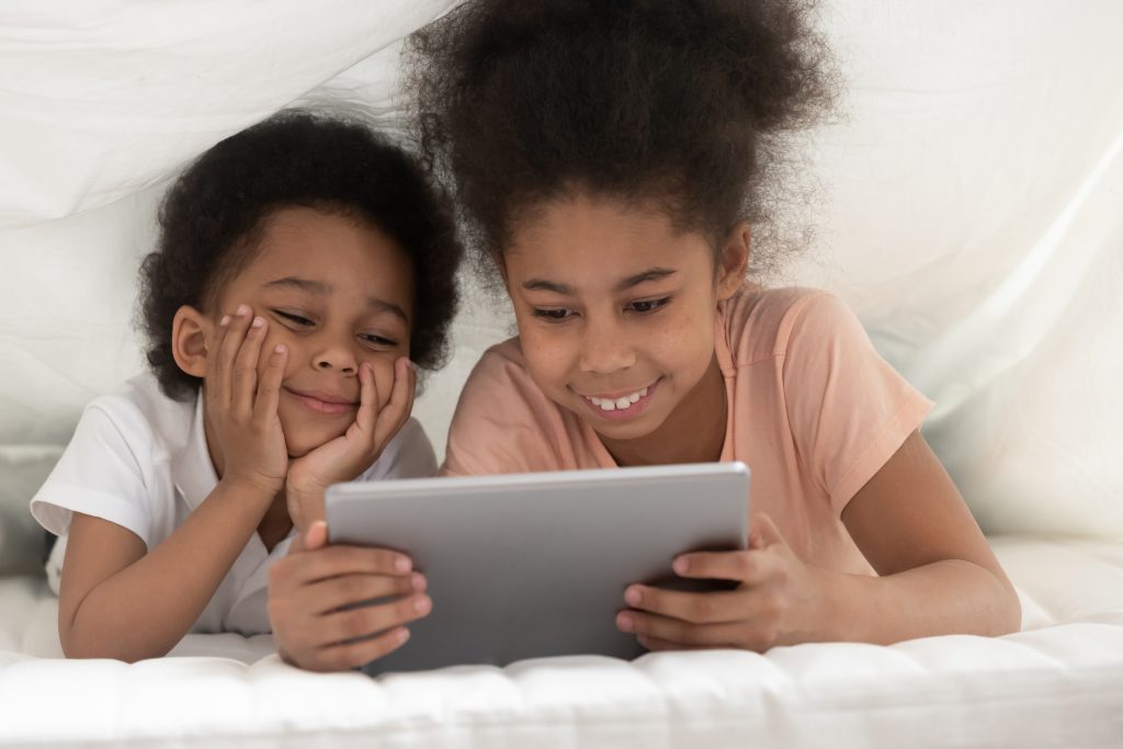 Smiling cute, young sister and brother lying under white blanket watching tablet together.