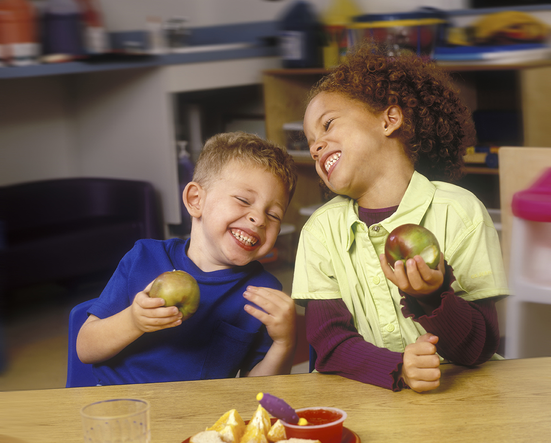 A young caucasian boy and an brown-skinned girl eating apples at a table and laughing wildely.