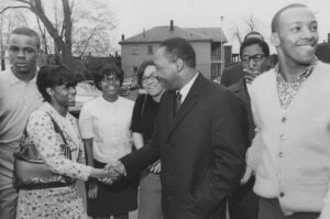 The photograph depicts Reverend Martin Luther King, Jr. outside Glenville High School, shaking hands with Sherida Foster of student council.