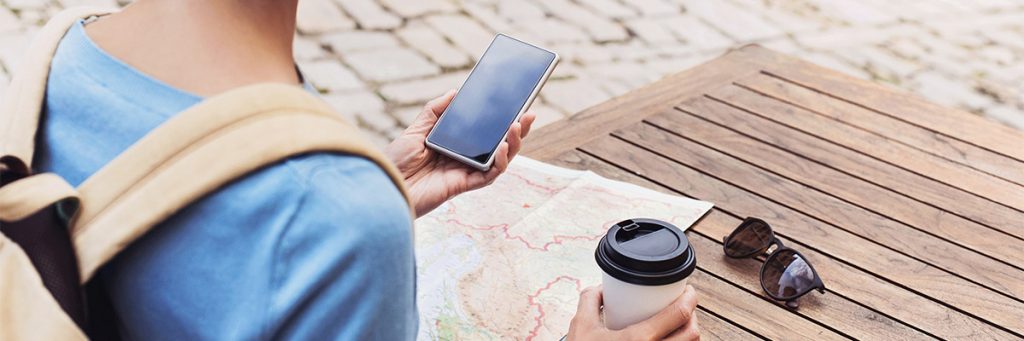 Picture of a young traveler looking at her cellphone while holding a cup of coffee. A map and a pair of sunglasses are on the table in front of her.